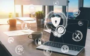 A laptop on a desk with coffee cups, documents, and office supplies, overlaid with digital cybersecurity icons like a lock and shield, reflecting 2020 cybersecurity trends in data protection and IT security within an office environment.